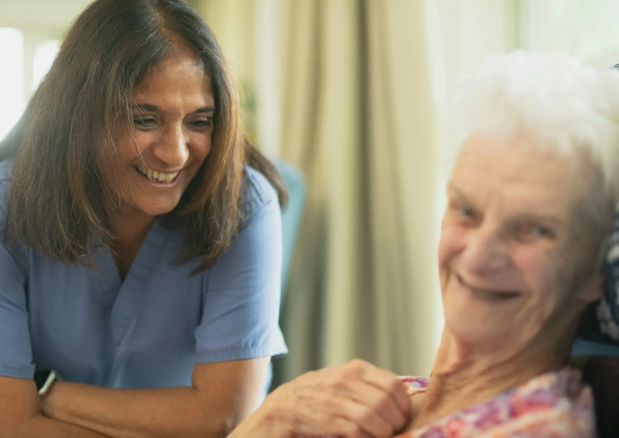 A smiling caregiver sits close to a laughing older woman