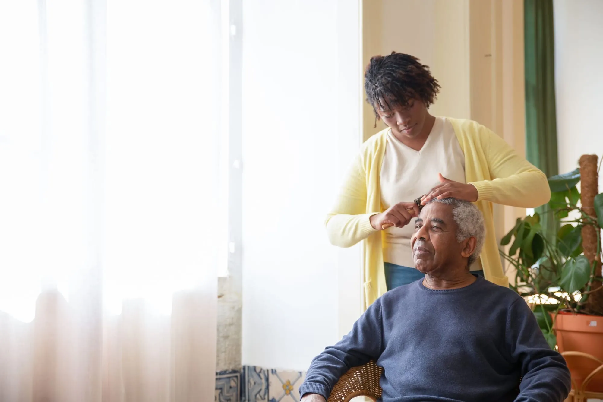 A caregiver gently combs an older man’s hair while he sits in a sunlit room