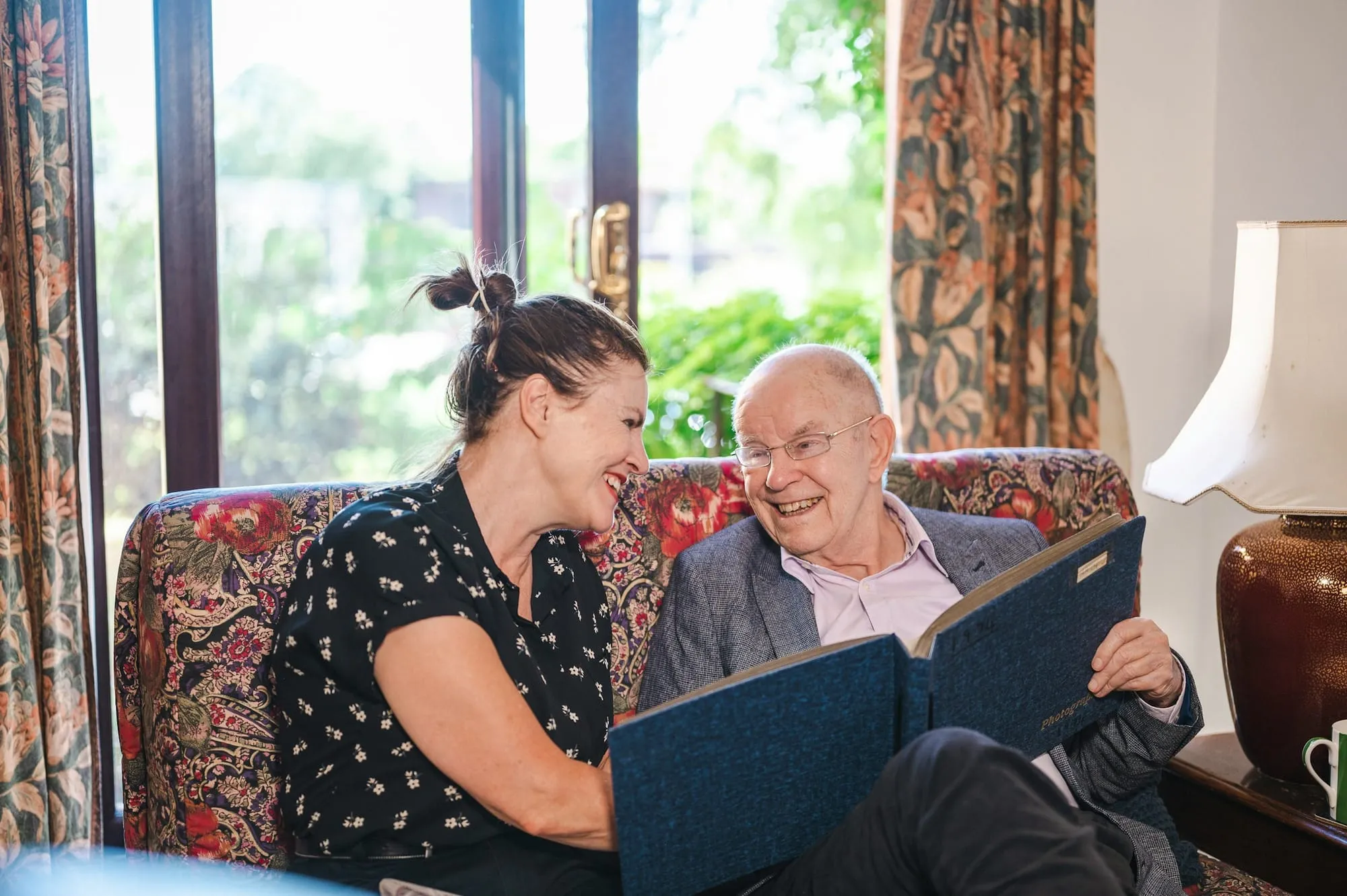 A woman and an older man laugh together over a photo album on a patterned sofa