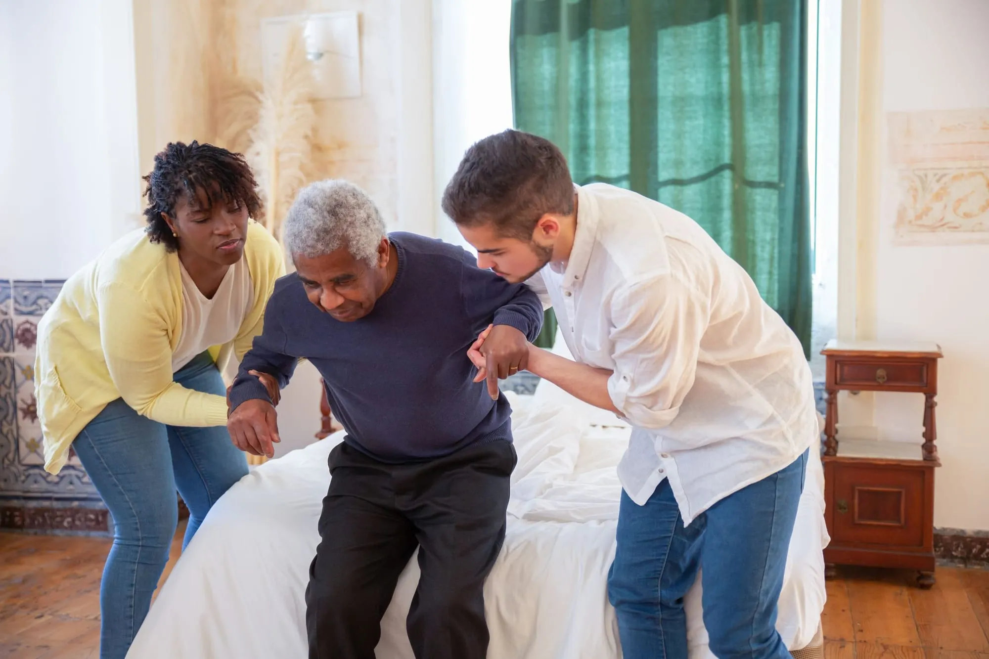 Two caregivers help an older man rise from the edge of his bed