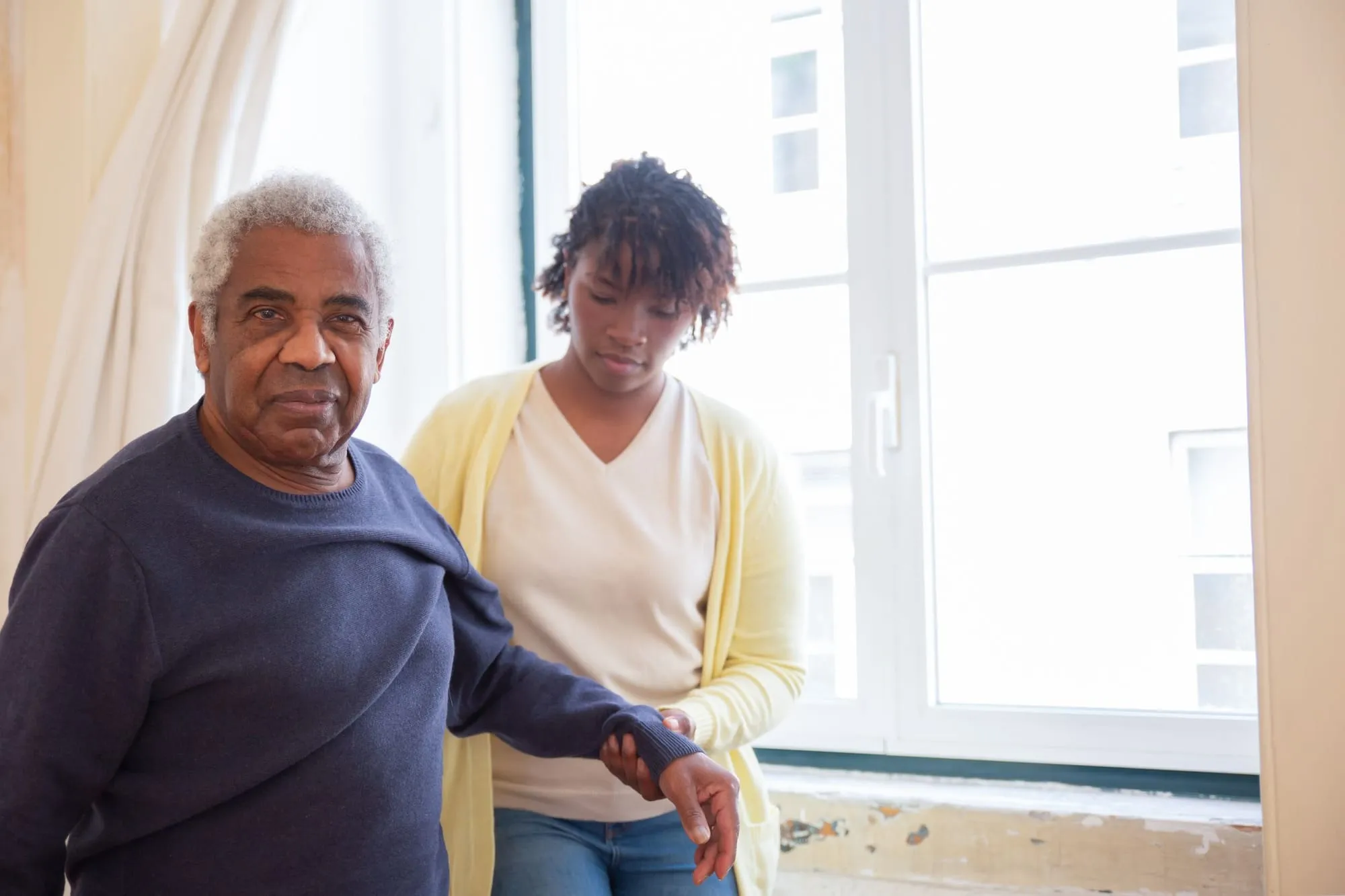 A caregiver steadies an older man’s arm as he stands by a window
